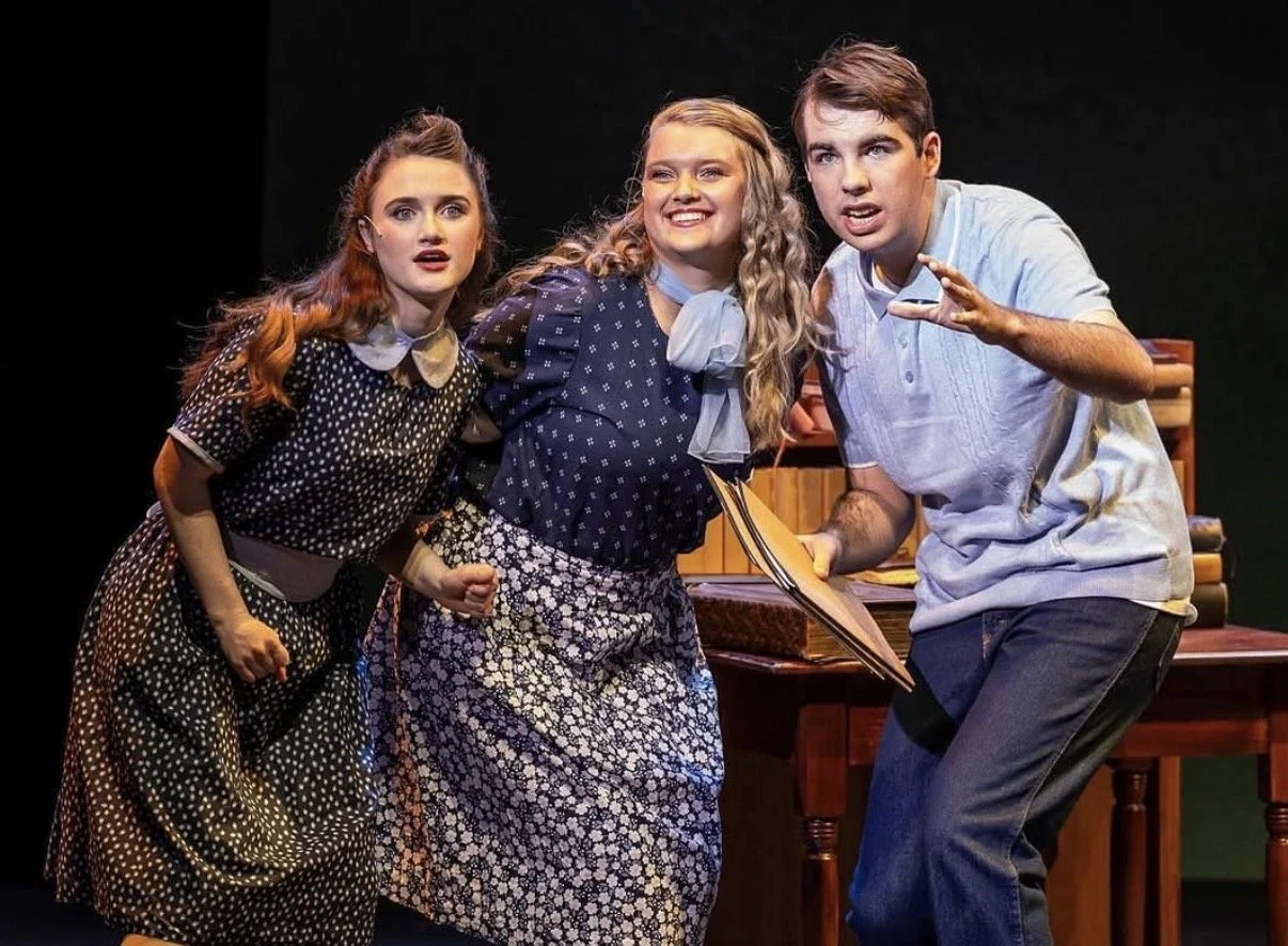 Three young actors on stage, dressed in vintage clothing, appear to be performing a scene with surprised and expressive facial expressions. The scene includes wooden furniture and a book, suggesting a period or school setting.
