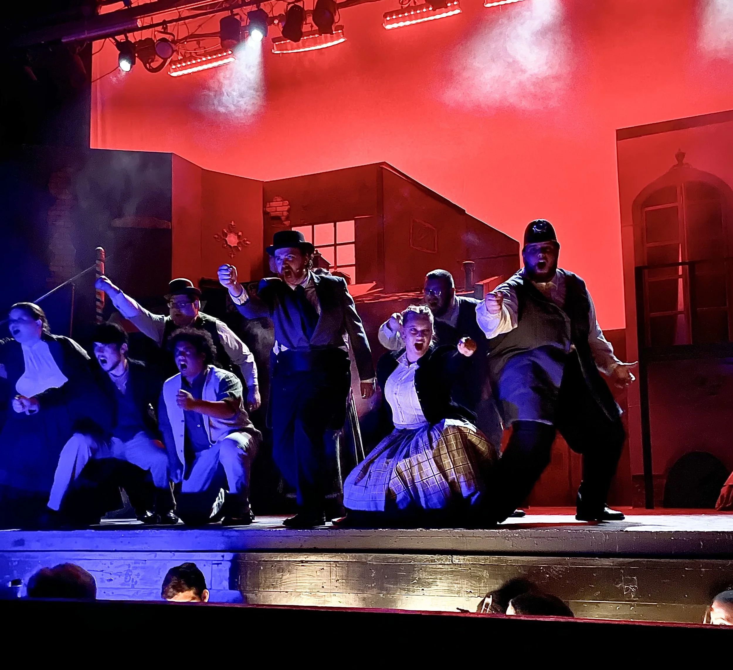 Stage scene with actors performing in costume, under red and blue lighting, in front of a set with windows and doors, with audience members visible at the bottom.