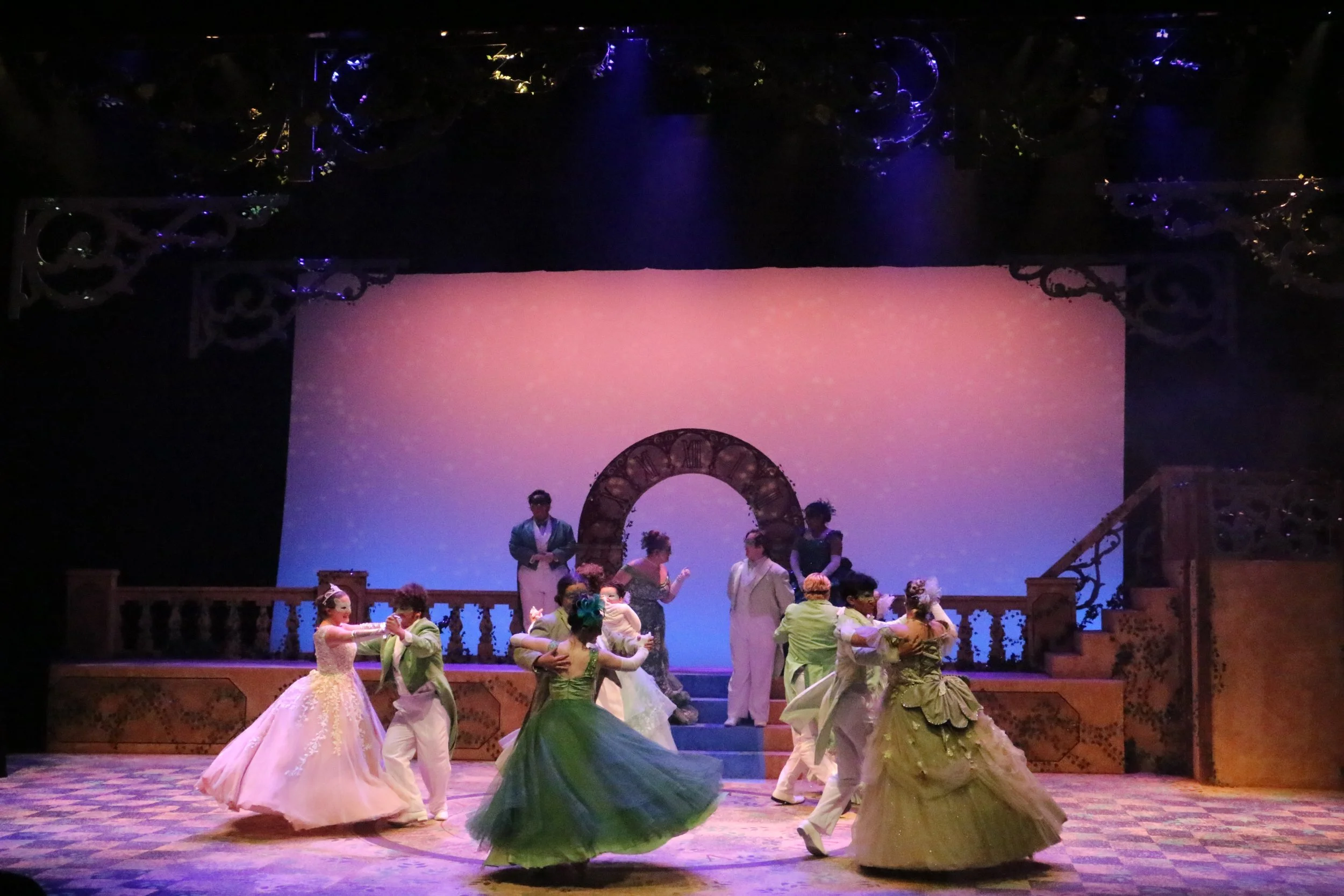 A stage performance with people dressed in elegant period costumes, including ball gowns and formal attire, engaging in a dance or scene from a theatrical production. The background features a light pink and purple gradient with decorative elements, and there are performers on the stage.