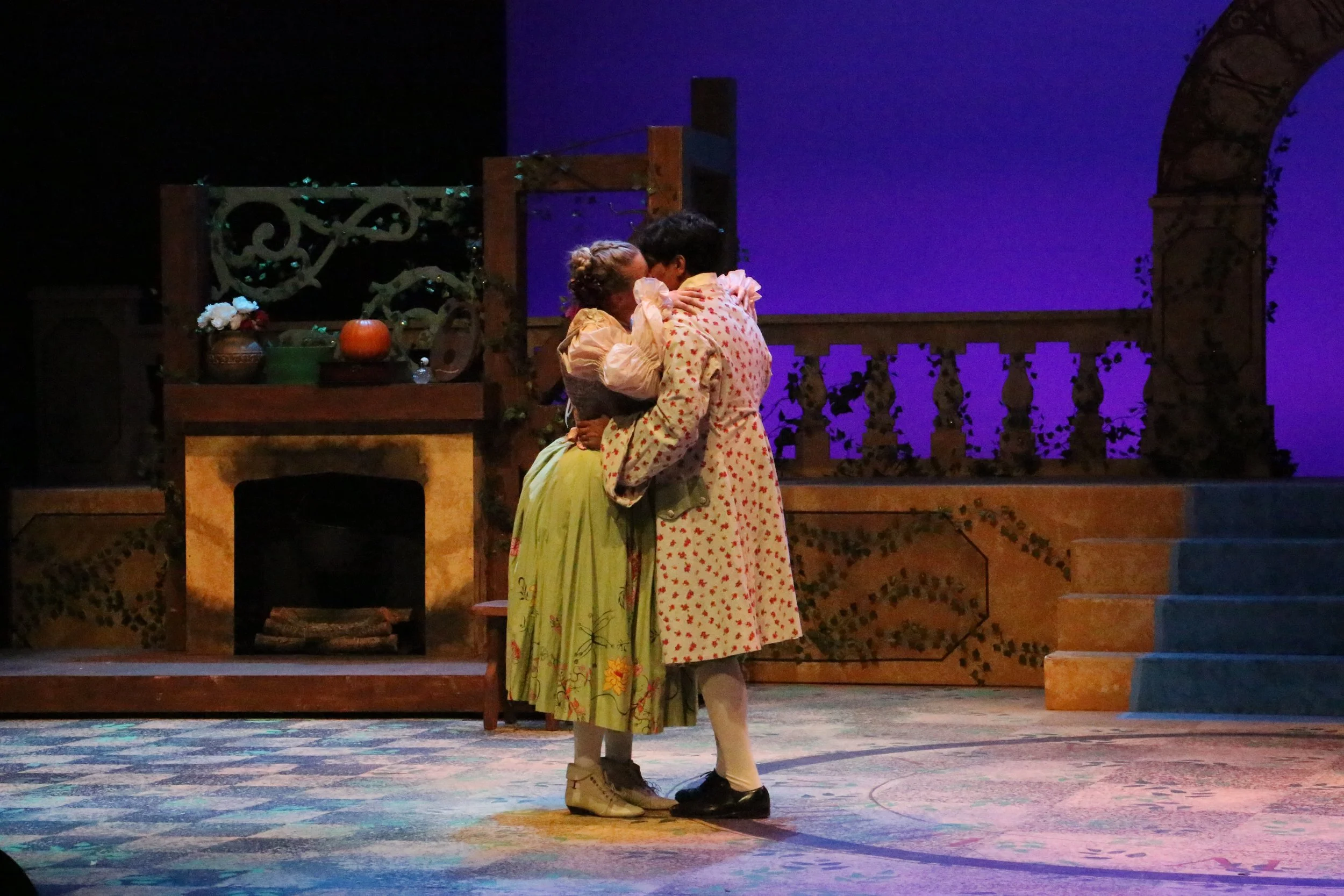 Two actresses in vintage costumes embracing on stage with a scenic set of a porch, pumpkins, and flowers, lit with theatrical lighting.