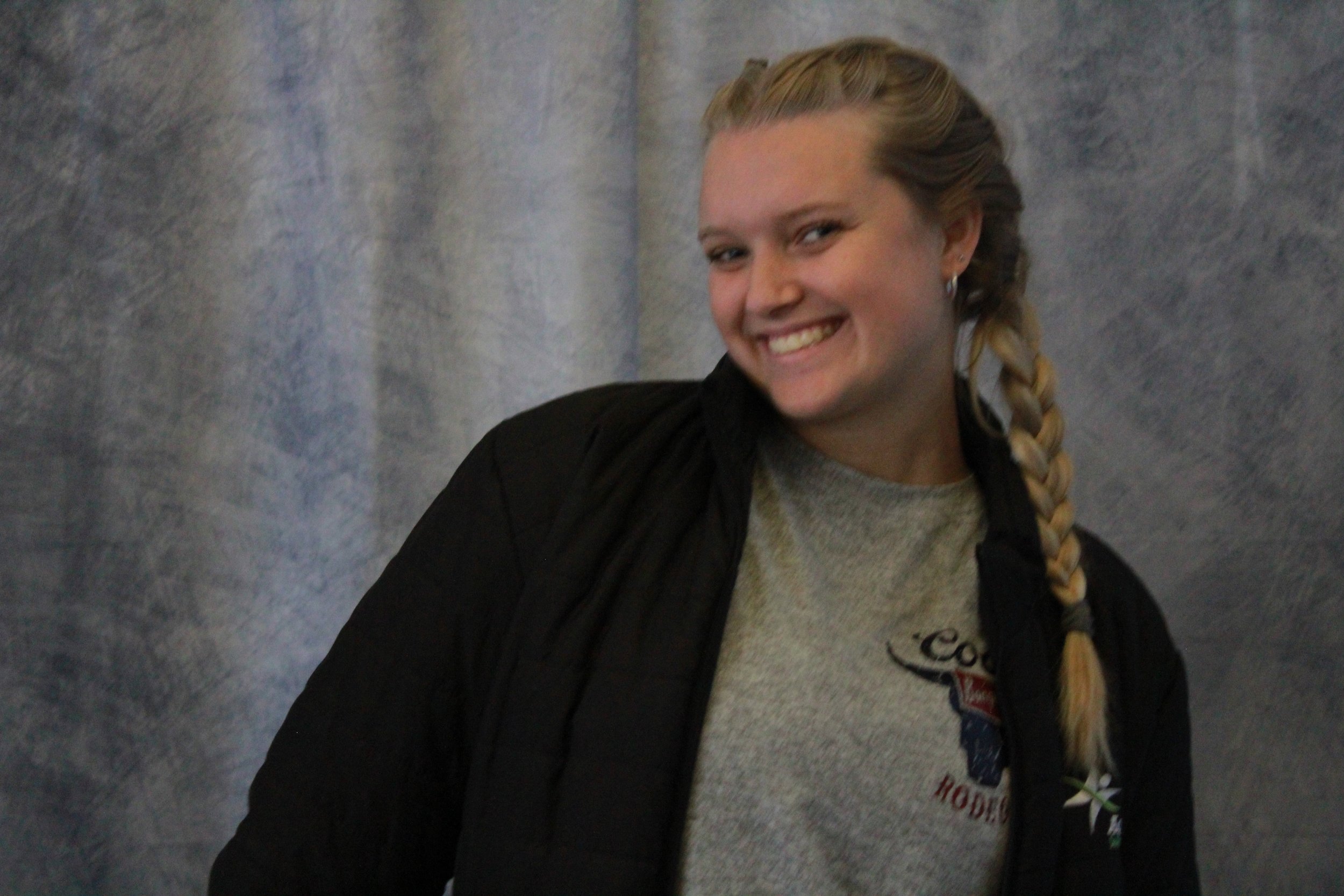 A young woman with blonde hair braided over her shoulder, smiling and wearing a gray shirt and a black jacket, standing against a gray textured wall.