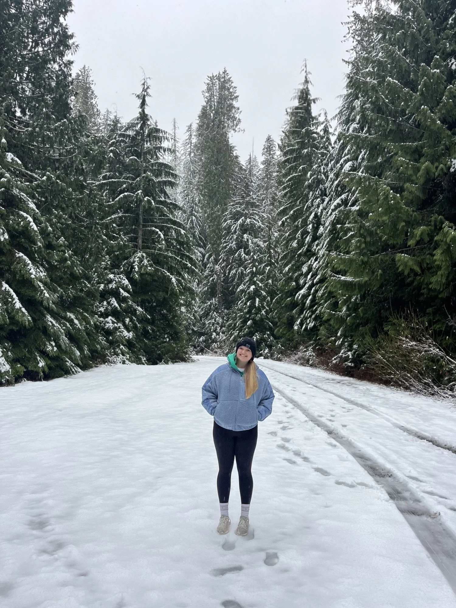 A smiling woman in a blue jacket and black leggings stands on a snow-covered trail in a forest of tall evergreen trees dusted with snow.