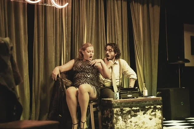 A woman and a man sitting on a stage with a green curtain behind them, performing a comedy act with a vintage microphone and sound equipment in front of them.