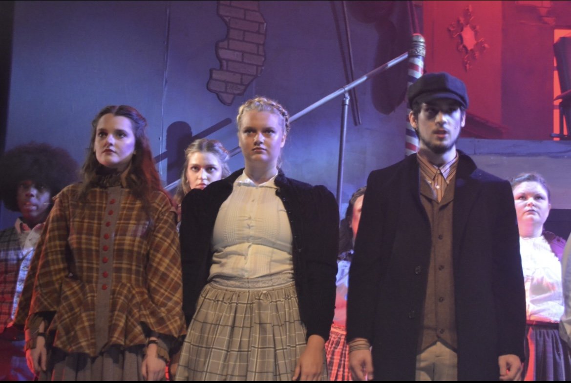 A group of young people dressed in period costumes on stage during a theatrical performance, with a dark background and theatrical lighting.