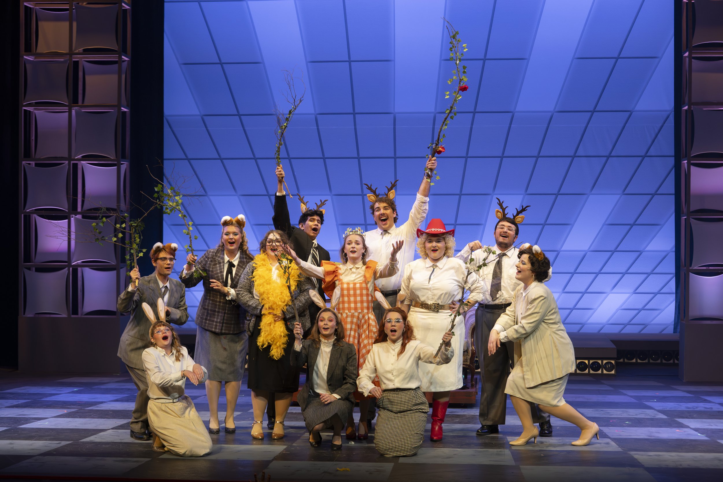 Group of actors on stage performing in a play, dressed in vintage costumes, some with reindeer antlers headbands, holding branches, under a blue-lit ceiling.
