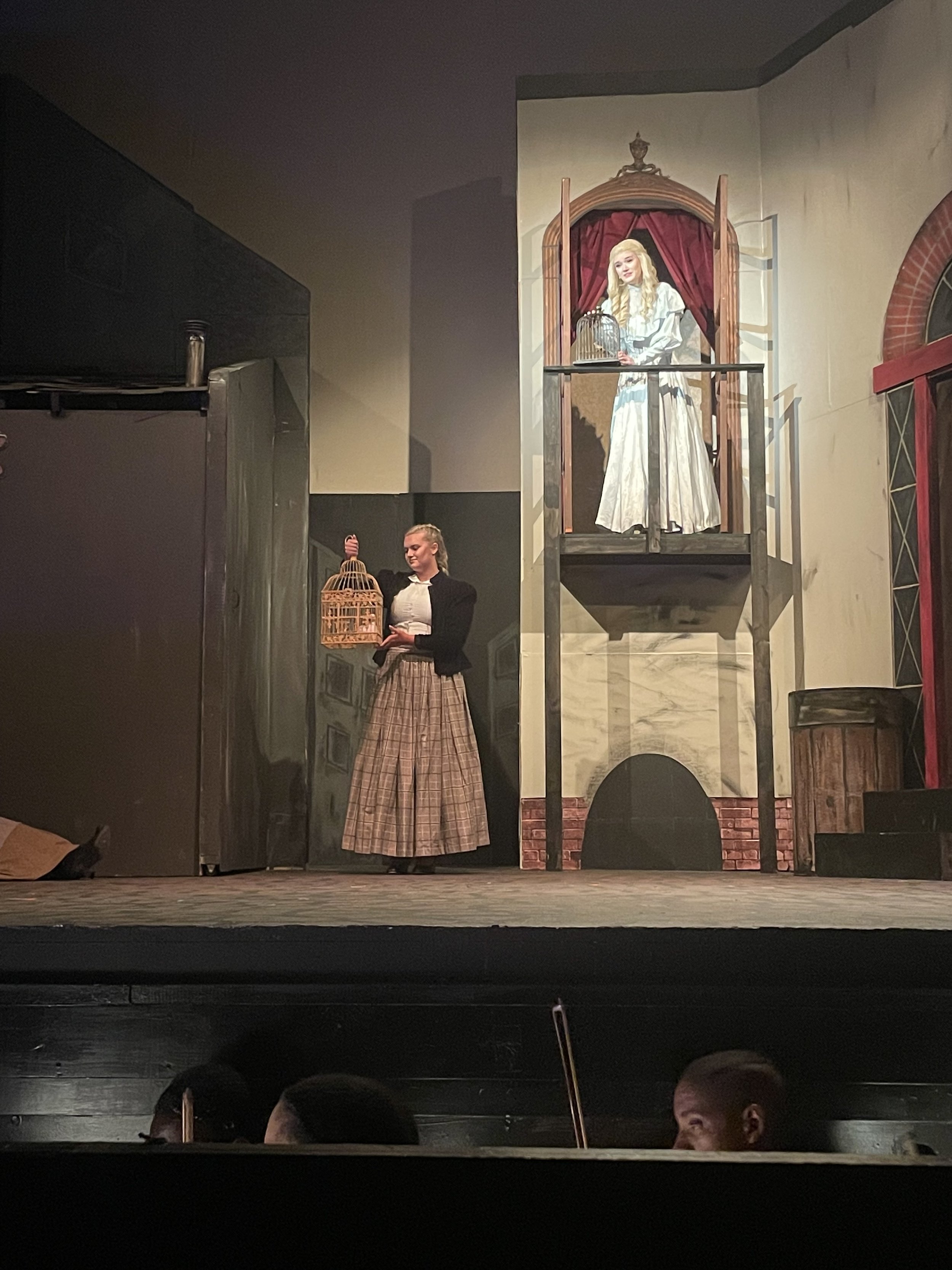 The image shows a theater stage with a scene featuring two women, one on a balcony and the other below holding a birdcage, acting as part of a play.