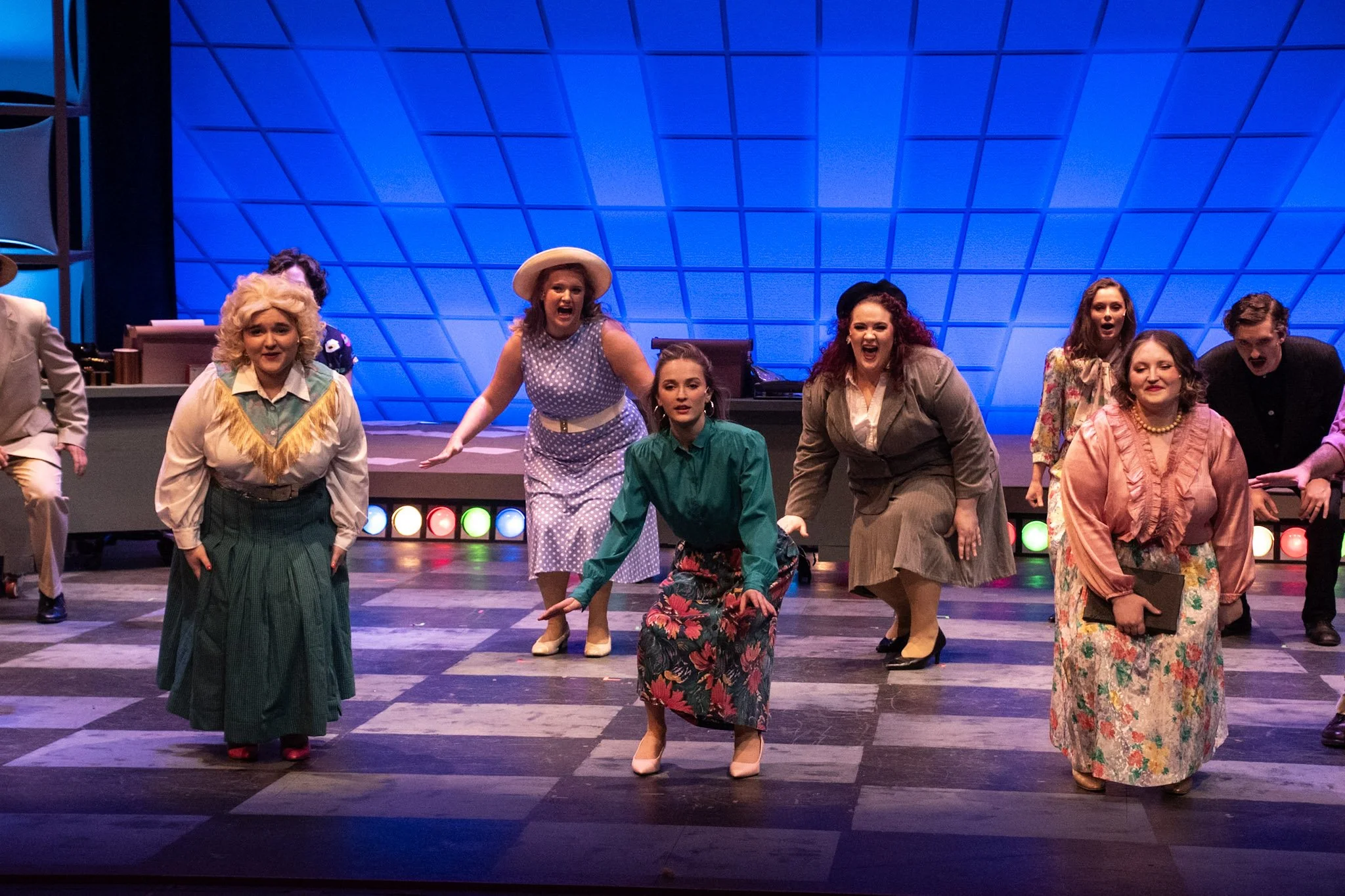 Actors performing on stage in costume, with a blue geometric backdrop and colorful stage lights.