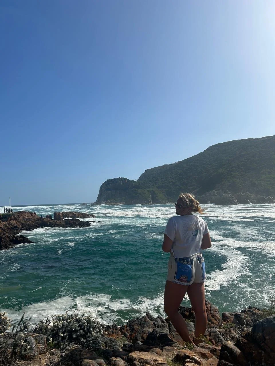 A woman with blonde hair wearing sunglasses, a white shirt, and shorts standing on rocks by the ocean, with waves and a green hillside in the background under a clear blue sky.