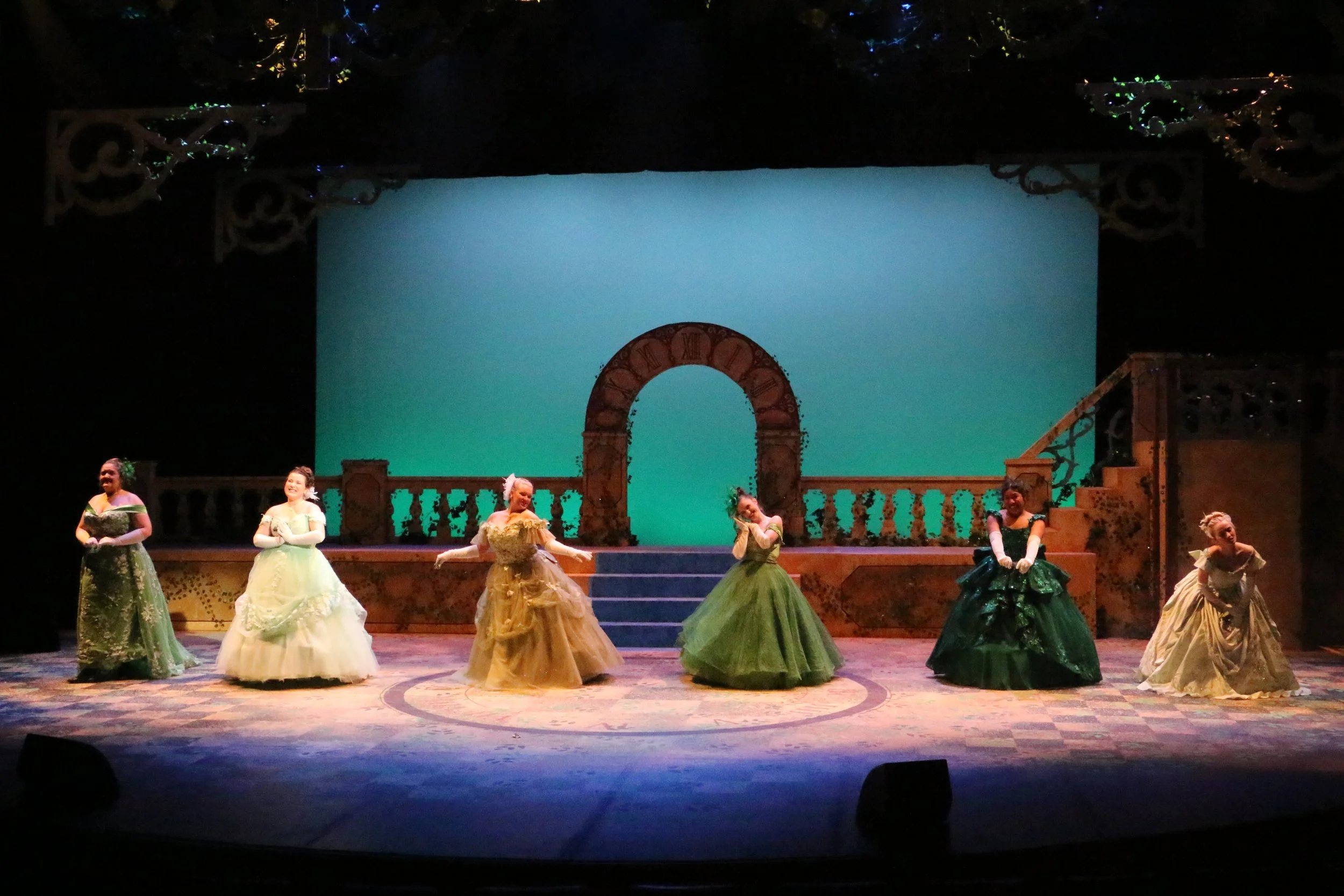 Stage scene from a ballet performance featuring women in colorful, elegant costumes with full skirts, standing in a semi-circle on a decorated stage with a large blue backdrop and ornate architectural props.