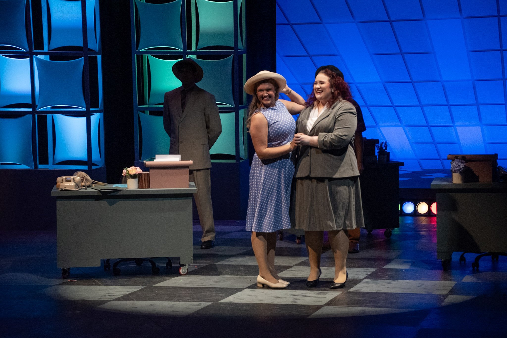 Two women in vintage-style clothing hugging on stage during a theatrical performance. One woman has curly hair and is wearing a gray blazer and skirt, while the other is wearing a blue polka-dot dress and a wide-brimmed hat. A man in a suit stands in the background to the left. The stage has a modern geometric blue backdrop, a desk with a rotary phone, a small flower arrangement, and some papers.