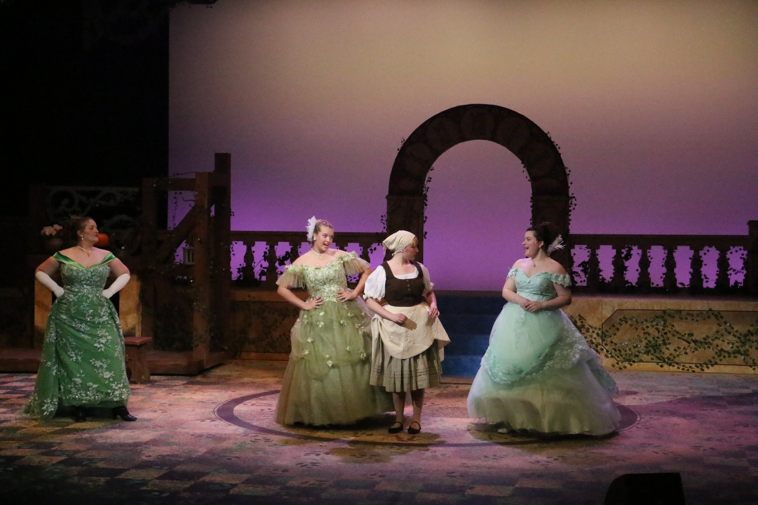 Women dressed in elaborate, period costumes are performing on a stage set resembling a historical scene with an arch and a castle wall in the background.