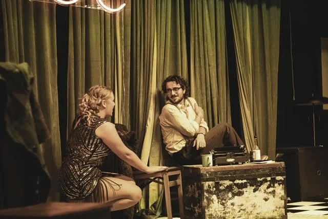 Two people sitting on stools and talking behind a green curtain backdrop at an indoor event or performance.