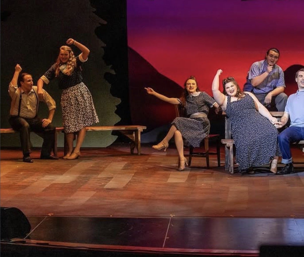 The image shows a theatrical stage with five actors in period costumes performing a scene. Four actors are seated on chairs, and one actor is standing, all engaging in expressive gestures.