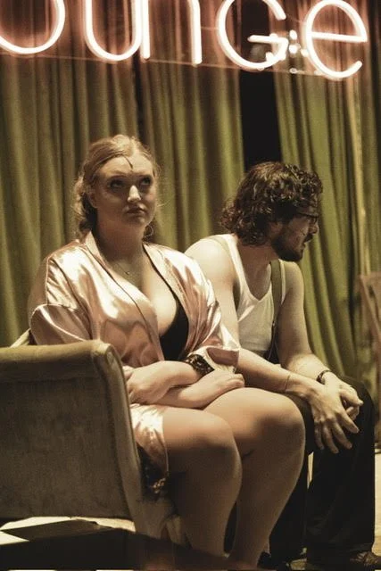 A woman and a man sitting on a stage with a neon sign that says 'courage' above them, with curtains in the background.