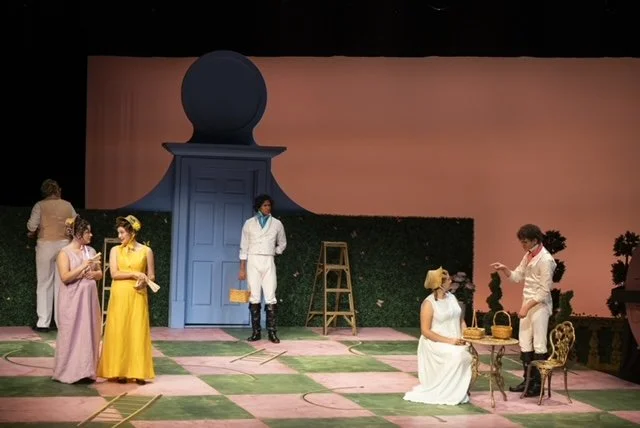 A stage play set with actors in costume, including a woman in a yellow dress, a woman in a pink dress, and two women in white clothing, with a blue door, ladder, and garden-like props on stage.