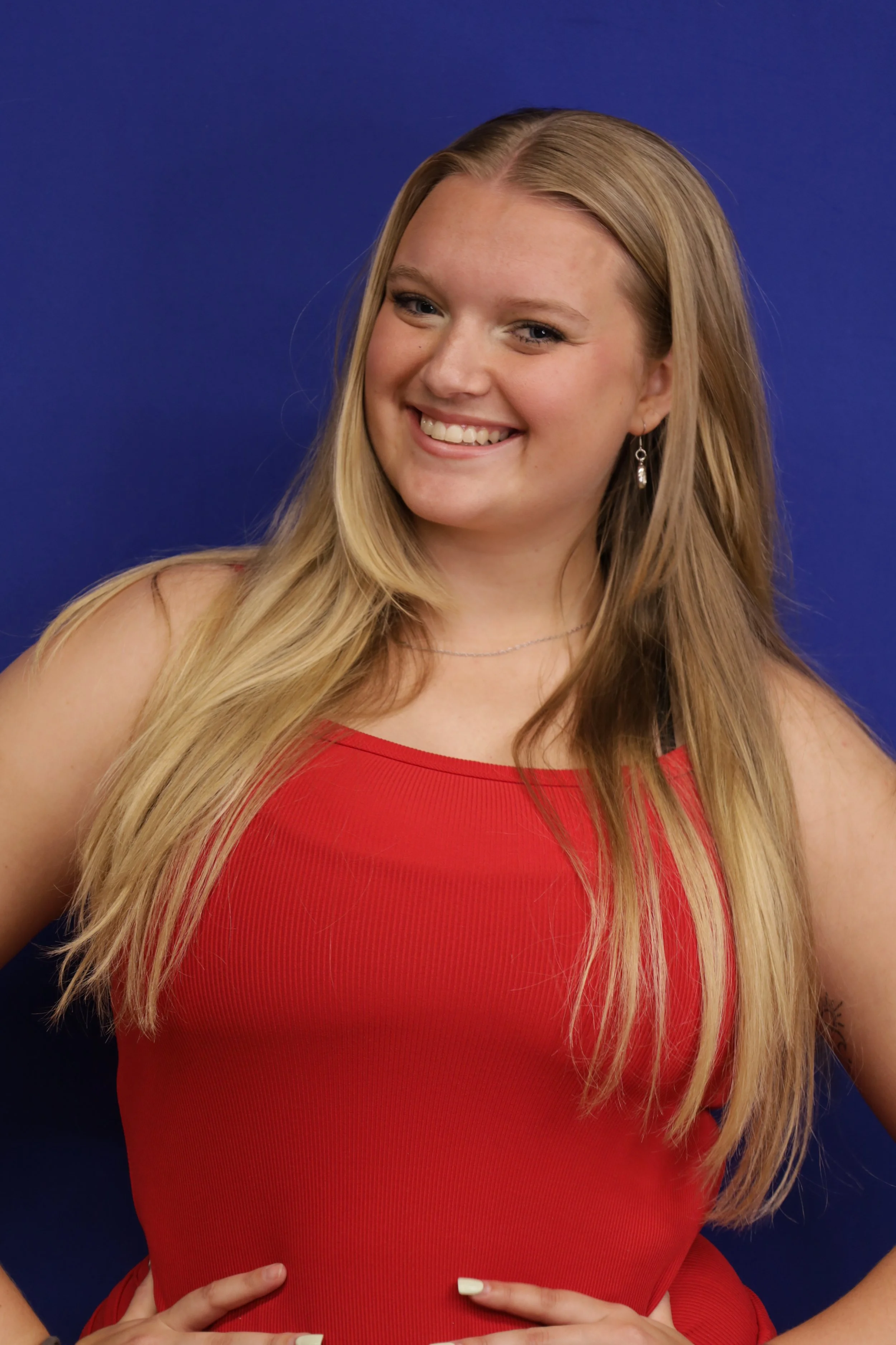 A woman with long blonde hair wearing a red sleeveless dress, smiling, against a blue background.