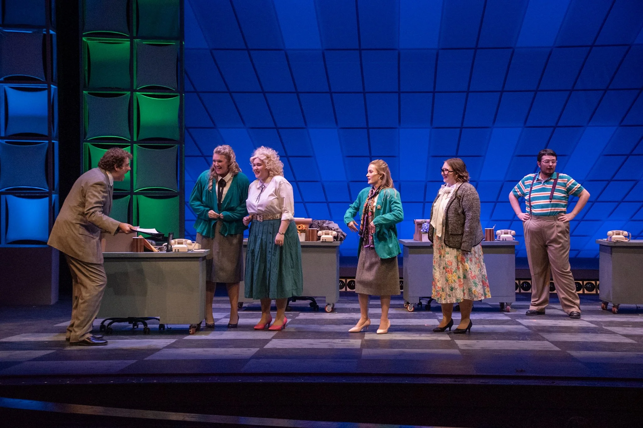 Group of six actors on stage in a theatrical performance, with five women and one man, wearing vintage clothing, engaged in a lively scene, with colorful set design in the background.