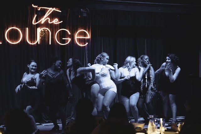 Seven women standing on a stage in front of a dark curtain, with a neon sign that reads 'The Lounge', in a nightclub or comedy club setting.