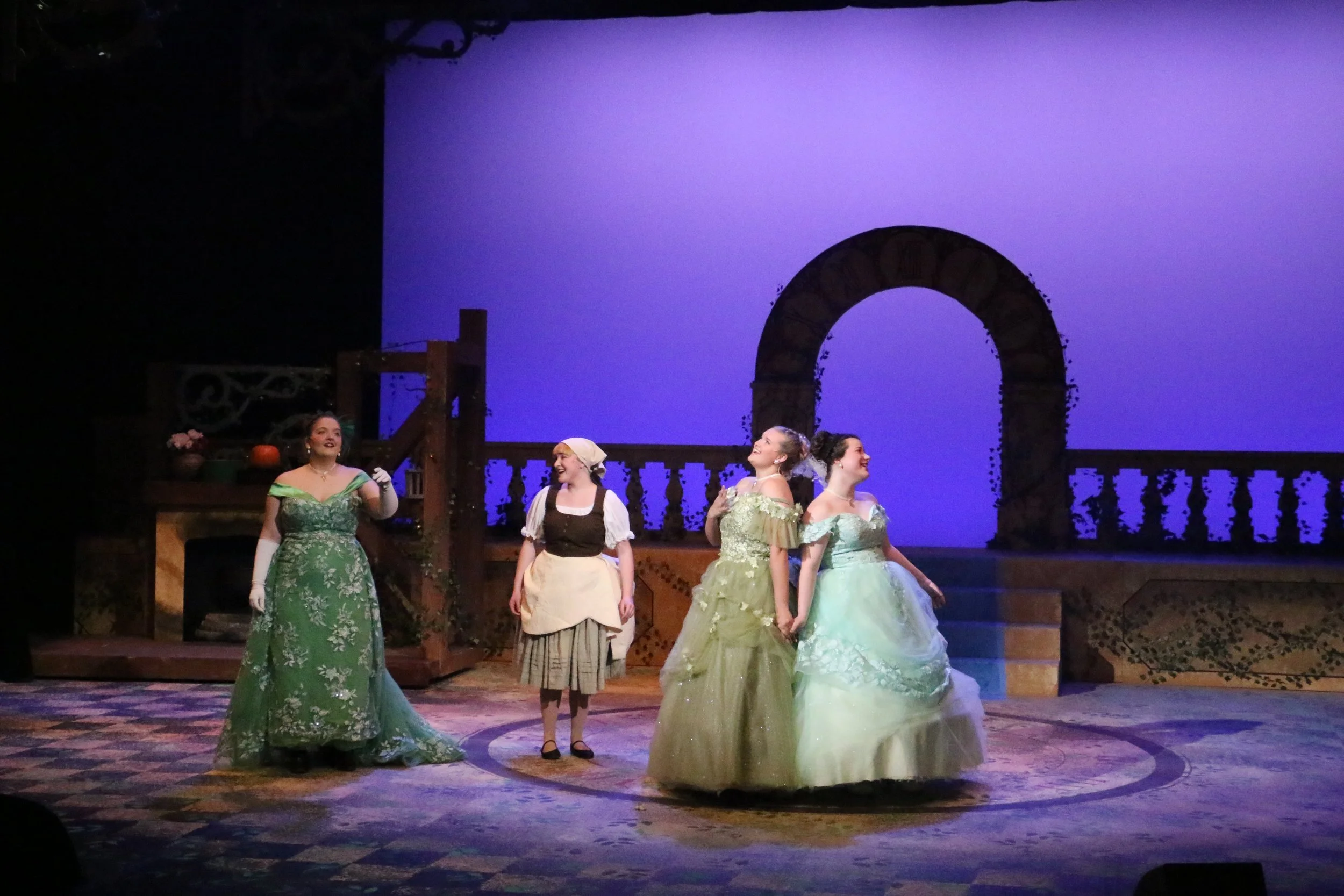 The image shows a theatrical stage with four women dressed in historical costumes, likely from the 18th or 19th century, performing in a scene. The set features a stone arch and a wooden balcony with pumpkins and flowers, illuminated with soft, warm lighting against a dark background.