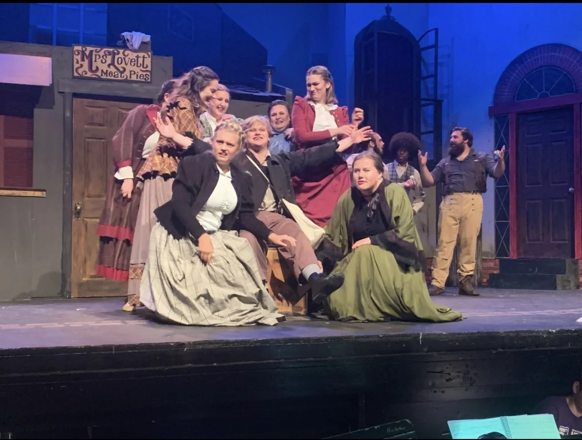 Actors performing on stage in a theatrical play set in a historical or rural setting, with a group of characters engaged in a scene near a food stand sign reading 'Lovett Meat Pies'.