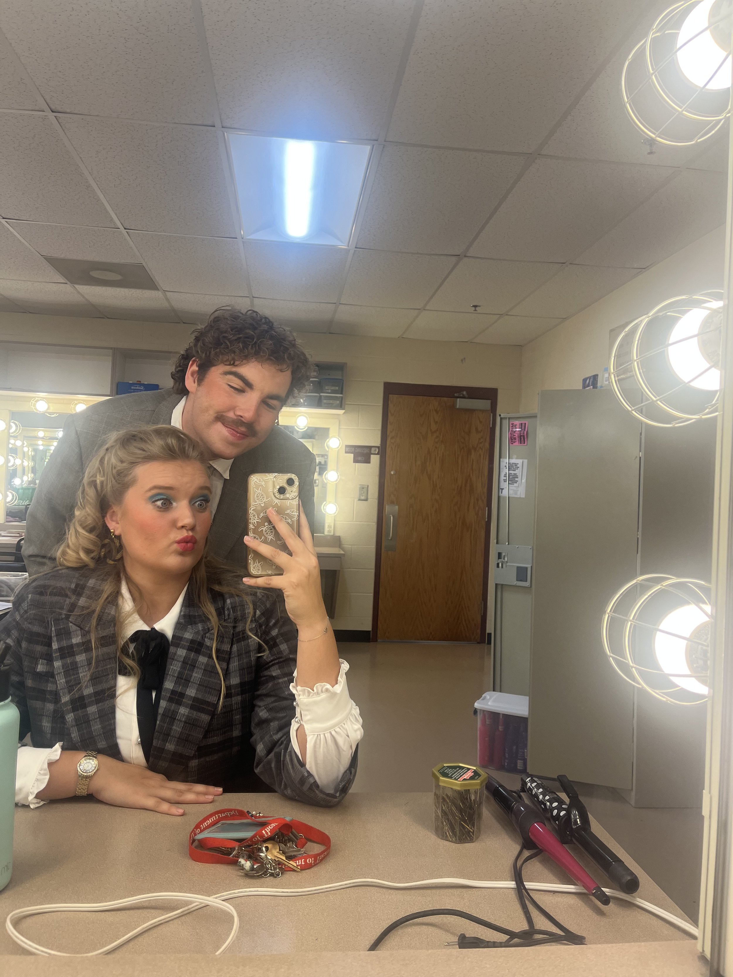Two people taking a selfie in a dressing room. The woman is in the foreground with makeup, and the man is behind her with curly hair and a mustache. The mirror shows their reflection, hair and makeup stations, and dressing room lights.
