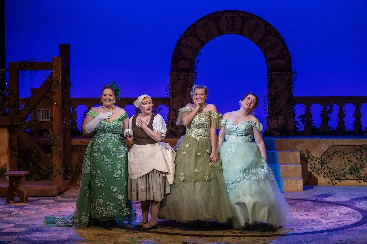 Four women dressed in period costumes on stage, smiling and holding hands, in front of a set with an arch and stairs, lit with blue and purple lighting.
