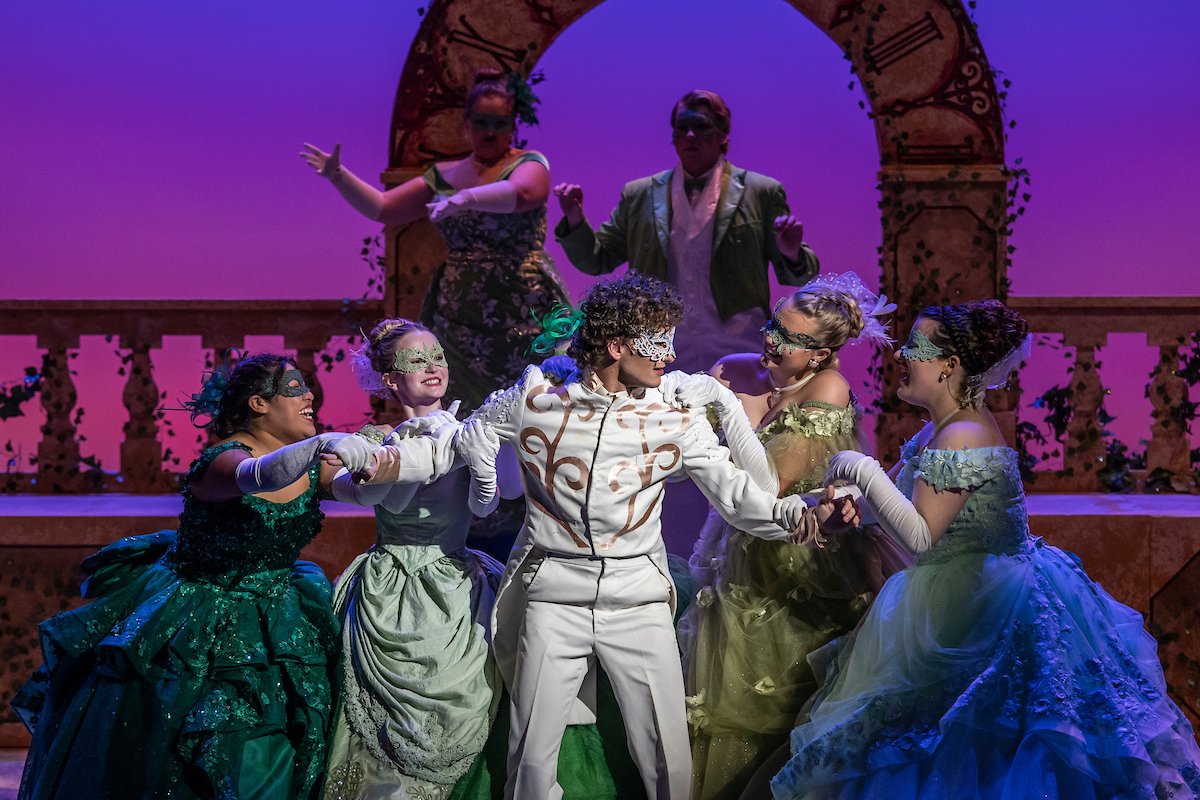 A theatrical stage play featuring six performers in elaborate costumes. The central figure is a woman in a white suit with gold embellishments, wearing a lace mask, surrounded by four women in colorful ball gowns and masks. Two more performers stand in the background, one with a green dress and the other with a jacket, under a decorative arch with Roman numerals.