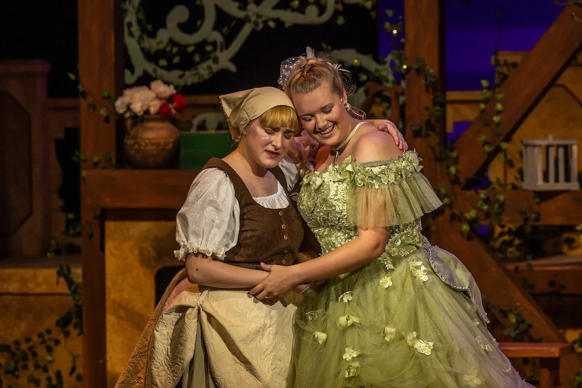 Two women in costume sharing an emotional moment on stage with a background of wooden structures and decorative plants.