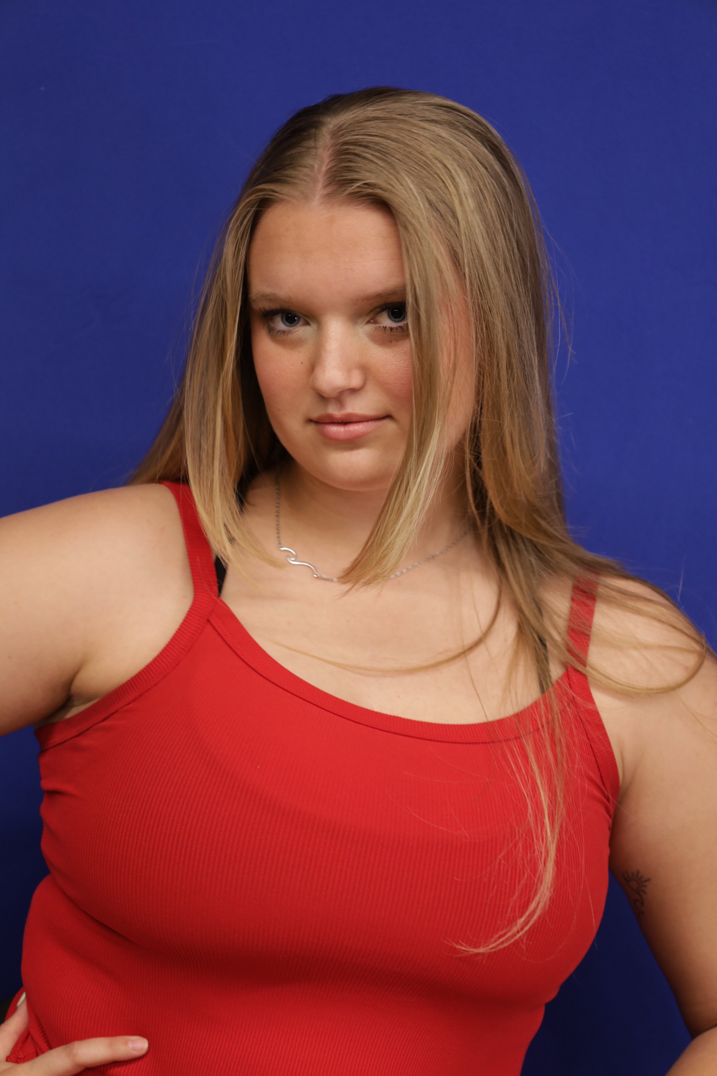 Young woman with blonde hair wearing a red tank top against a blue background.