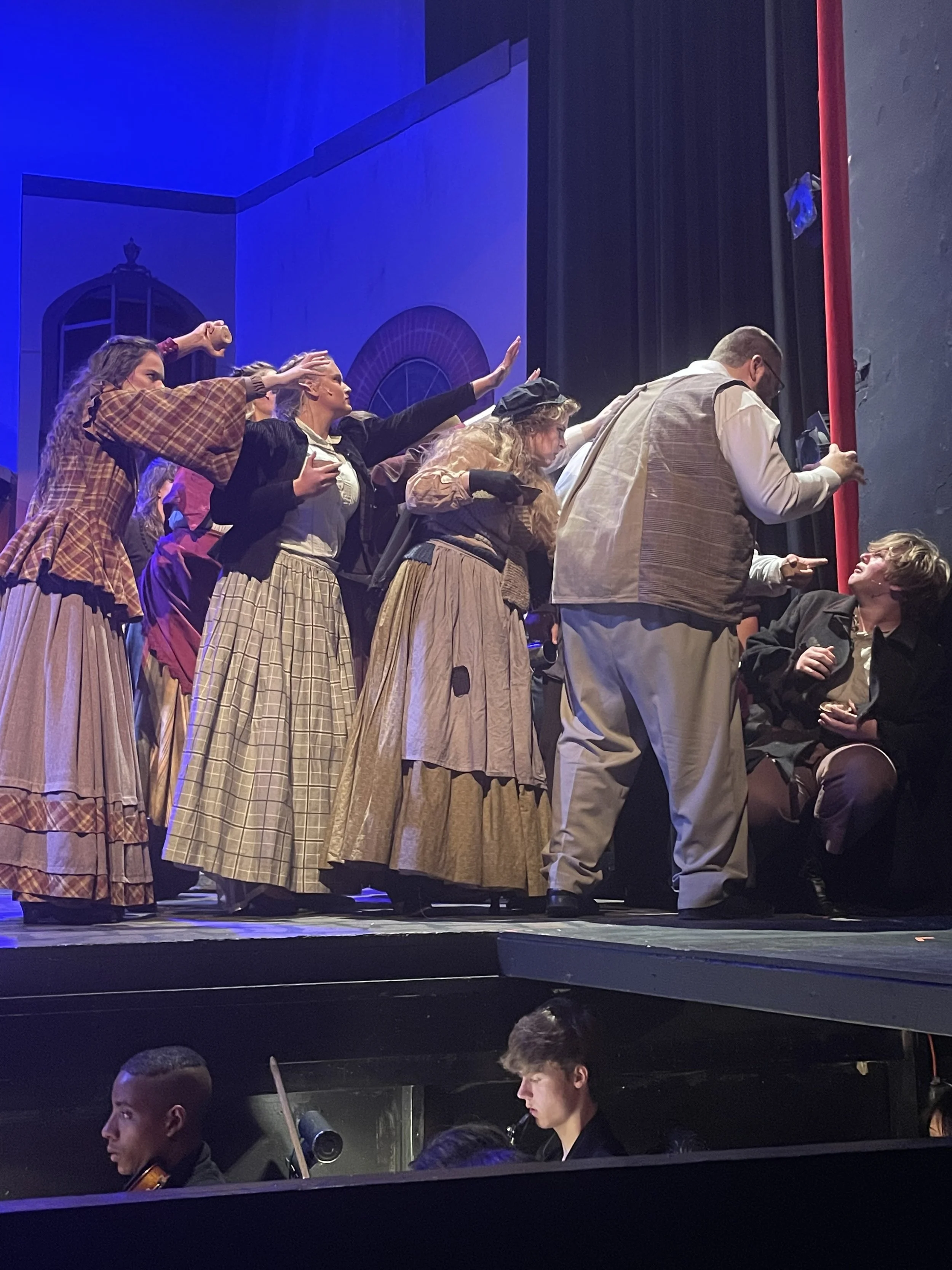 Stage scene with actors in period costumes performing a play, with musicians playing string instruments below the stage.