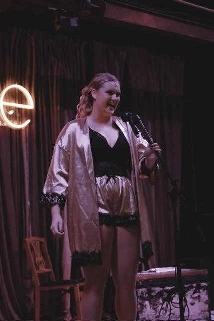 A woman performing on stage with a microphone, wearing a satin robe, shorts, and a black top, with a neon sign partially visible in the background.