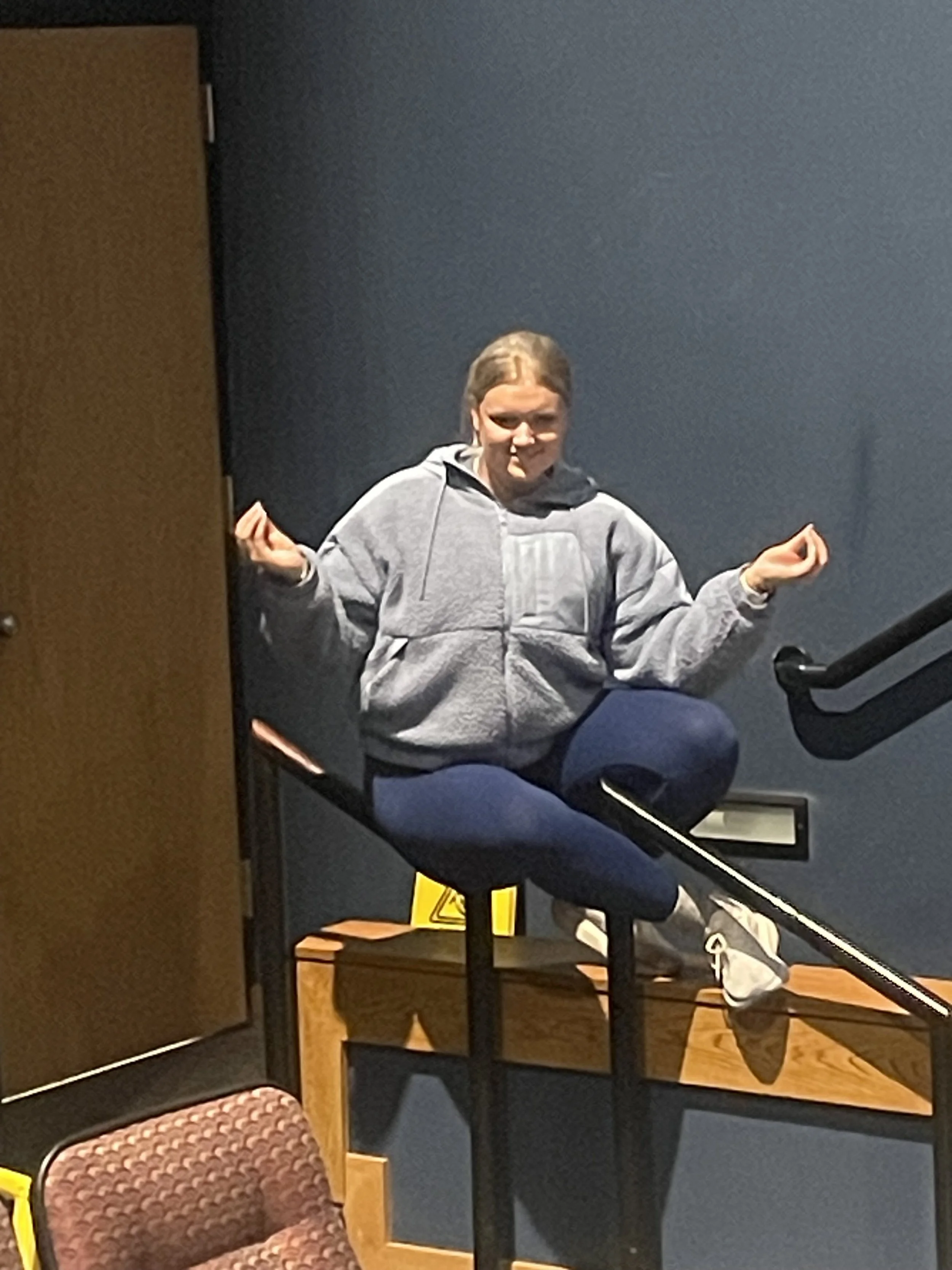 Person with long hair in a ponytail wearing a gray hoodie and jeans, making a funny face and gesturing with both hands, seated on a chair in a room with dark blue walls and various furniture.