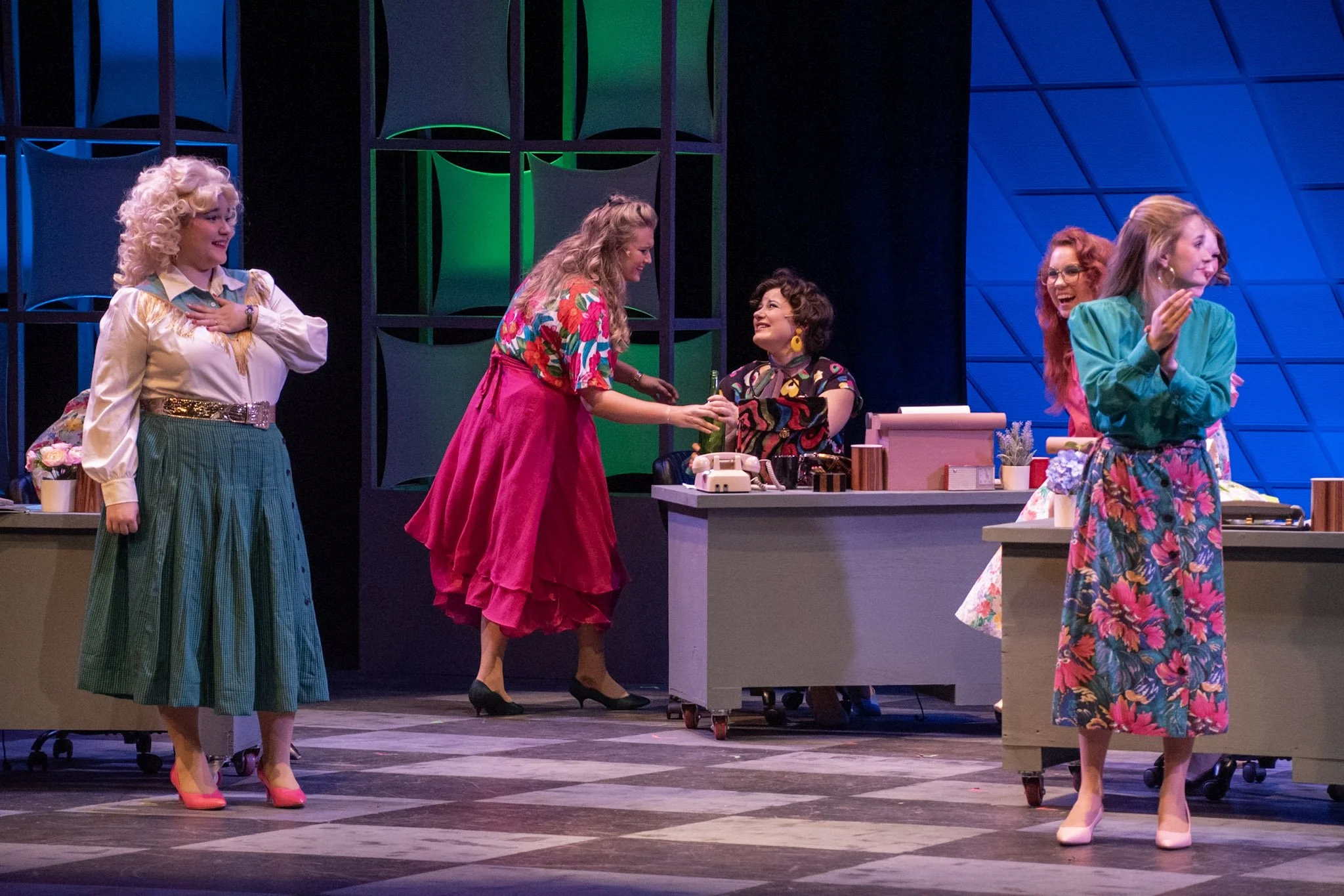 Four women dressed in colorful vintage clothing on a stage, engaging in a theatrical performance or rehearsal, with a woman in a floral blouse and pink skirt shaking hands with another woman in a bright skirt, while other women smile and laugh nearby.