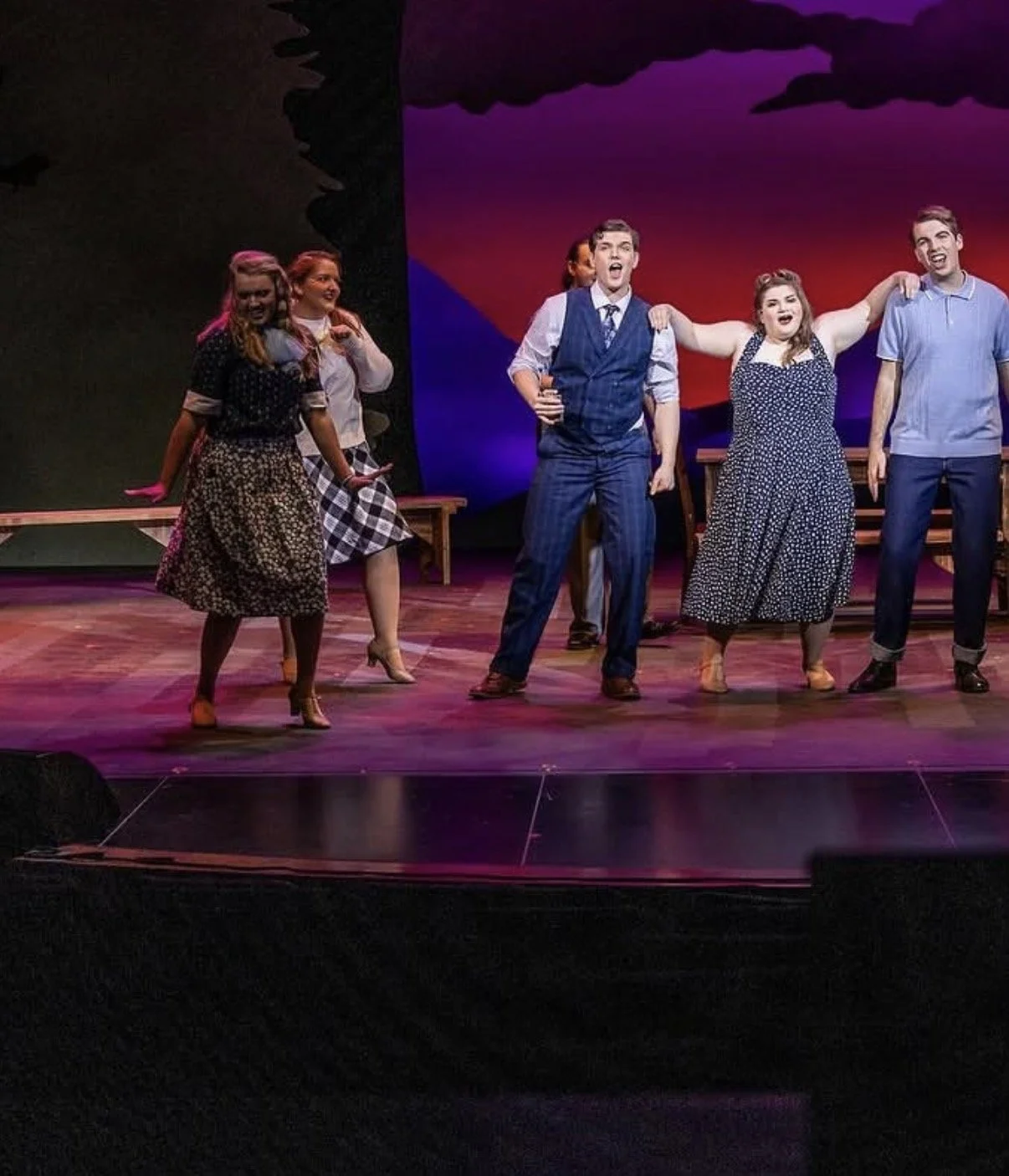 Actors performing a scene on stage. Four actors are standing in the foreground, two women and two men, wearing vintage-style clothing. Two more actors are visible in the background. The stage has wooden flooring and a colorful backdrop with purple and red hues.