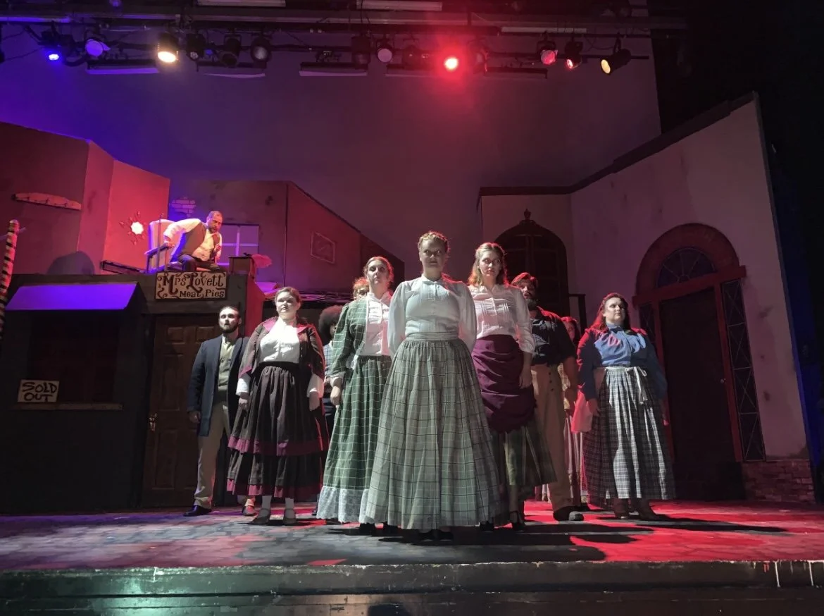The cast of a theatrical play onstage with actors standing in a line, some with serious expressions, under colorful stage lighting.