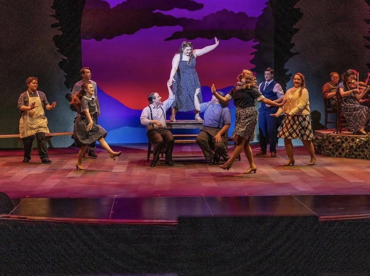Theater cast performs a musical scene with a woman in a polka dot dress being lifted by two men, while other cast members dance and play instruments on stage with a colorful sunset backdrop.