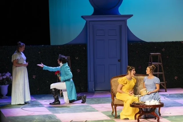 Two women in pastel-colored dresses sitting on a vintage sofa with a small table, and two women standing and kneeling on stage with a backdrop of a large blue door and butterfly decorations. A woman in a white dress stands near them, while the other woman in a blue dress kneels with an outstretched arm.