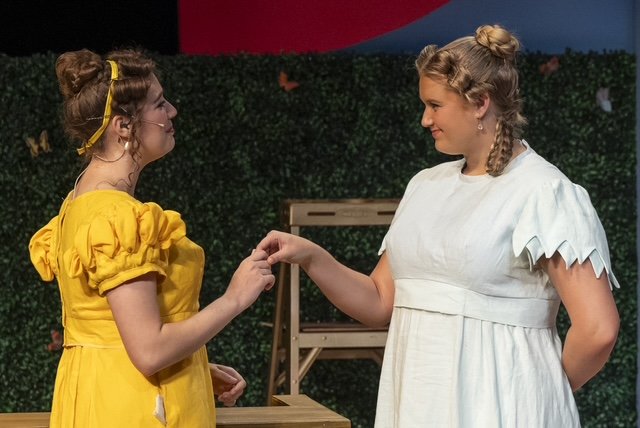 Two young women wearing costume dresses are holding hands and facing each other on stage.