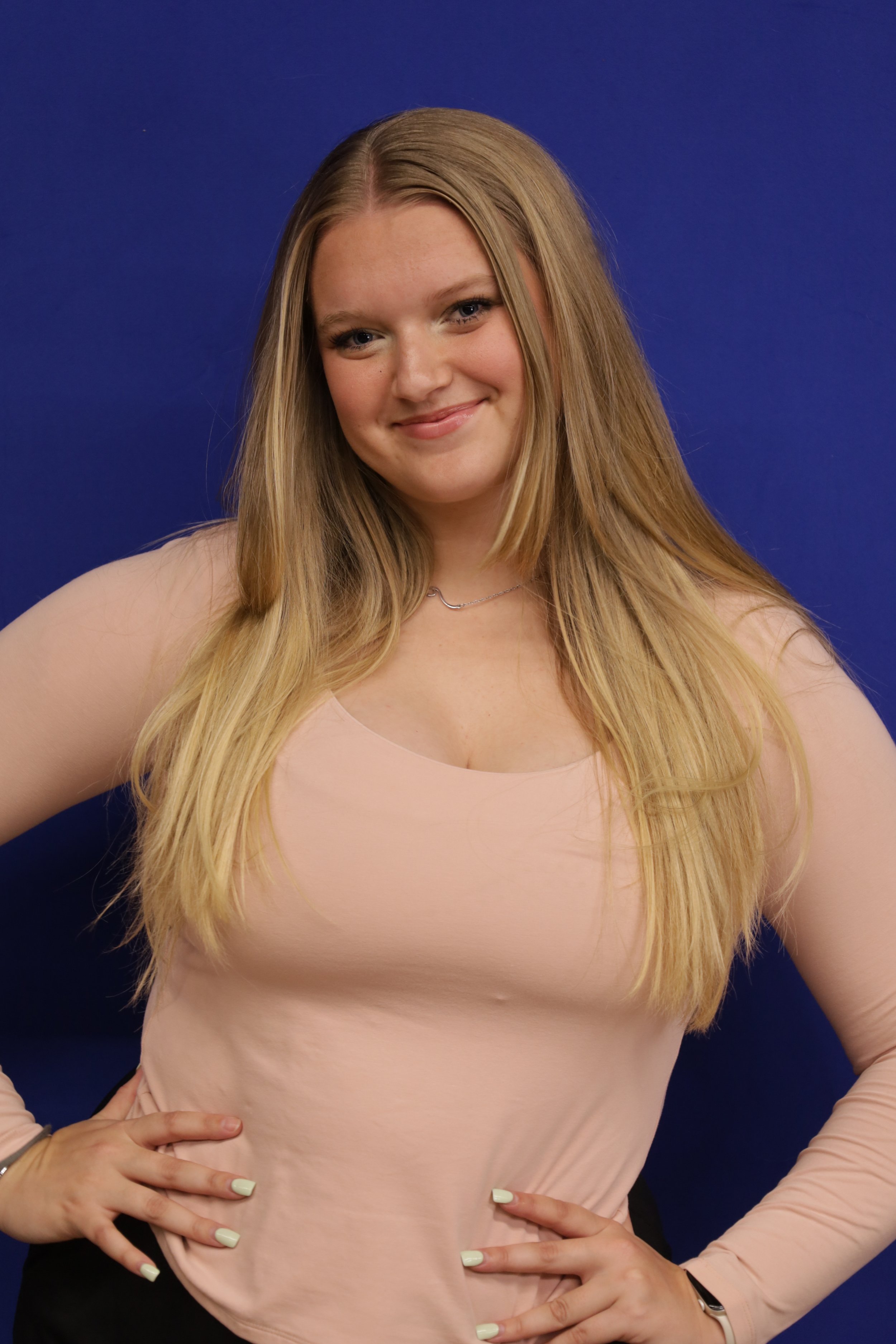 A woman with long blonde hair, wearing a light pink long-sleeved shirt and a silver necklace, standing against a blue background. She has a subtle smile, and her hands are placed on her hips.
