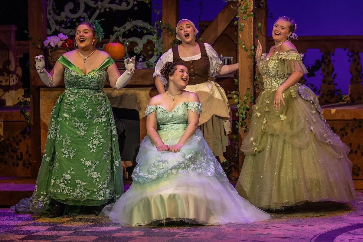 Four women in elaborate period costumes on stage, smiling and posing during a theatrical performance. The set background is decorated with flowers, pumpkins, and wooden beams.