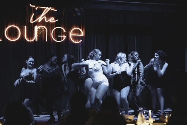 Women dancing on stage at a nightclub called The Lounge, with a neon sign in pink and white lights