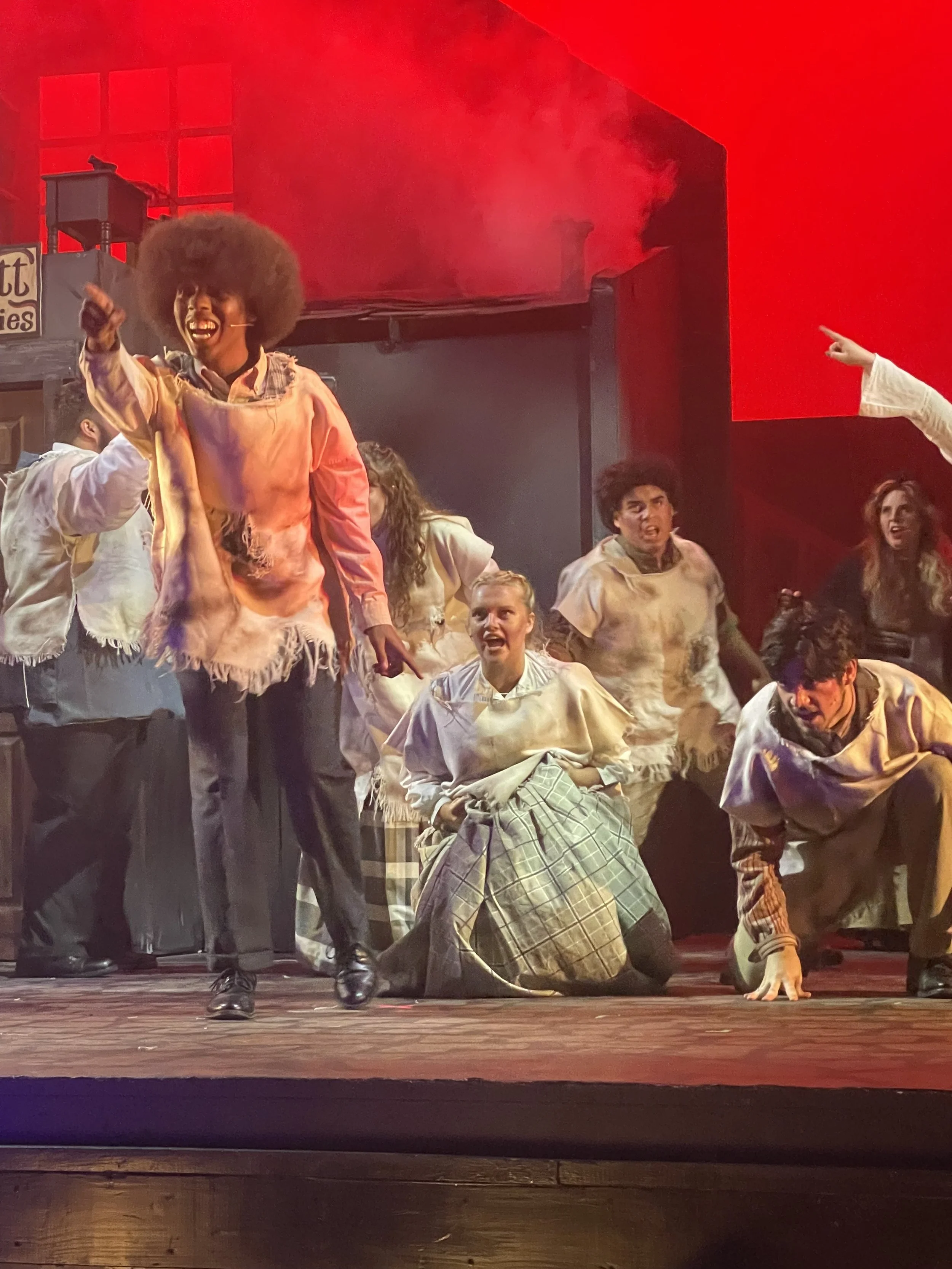 Actors perform a scene on stage with a red background, dressed in costumes, some with exaggerated facial expressions, and engaging in dramatic gestures.