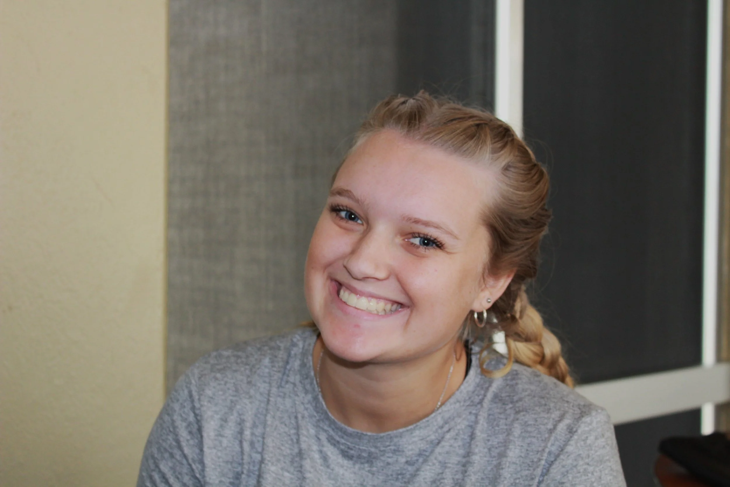 A young girl with long blonde hair tied in a braid, blonde eyebrows, light blue eyes, and a smile, wearing a grey top and earrings, sitting indoors near a dark sliding door.