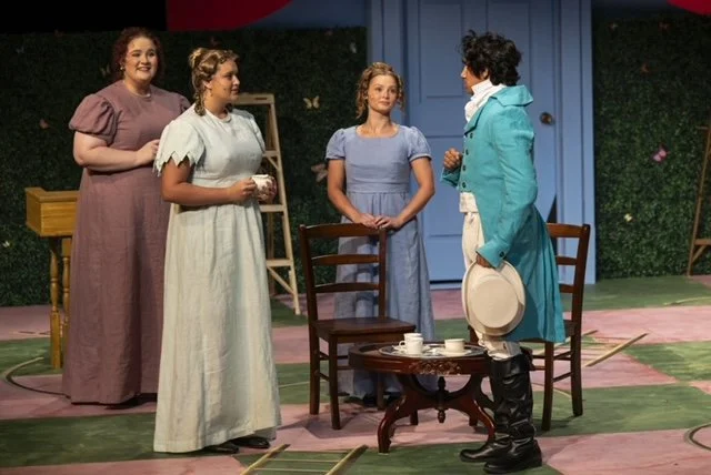 Four women are on a stage set that resembles a garden or backyard, with a blue door and green foliage. They are engaged in a conversation, with one woman in a bright blue coat and black shoes talking to three women in dresses, two of which are holding teacups.