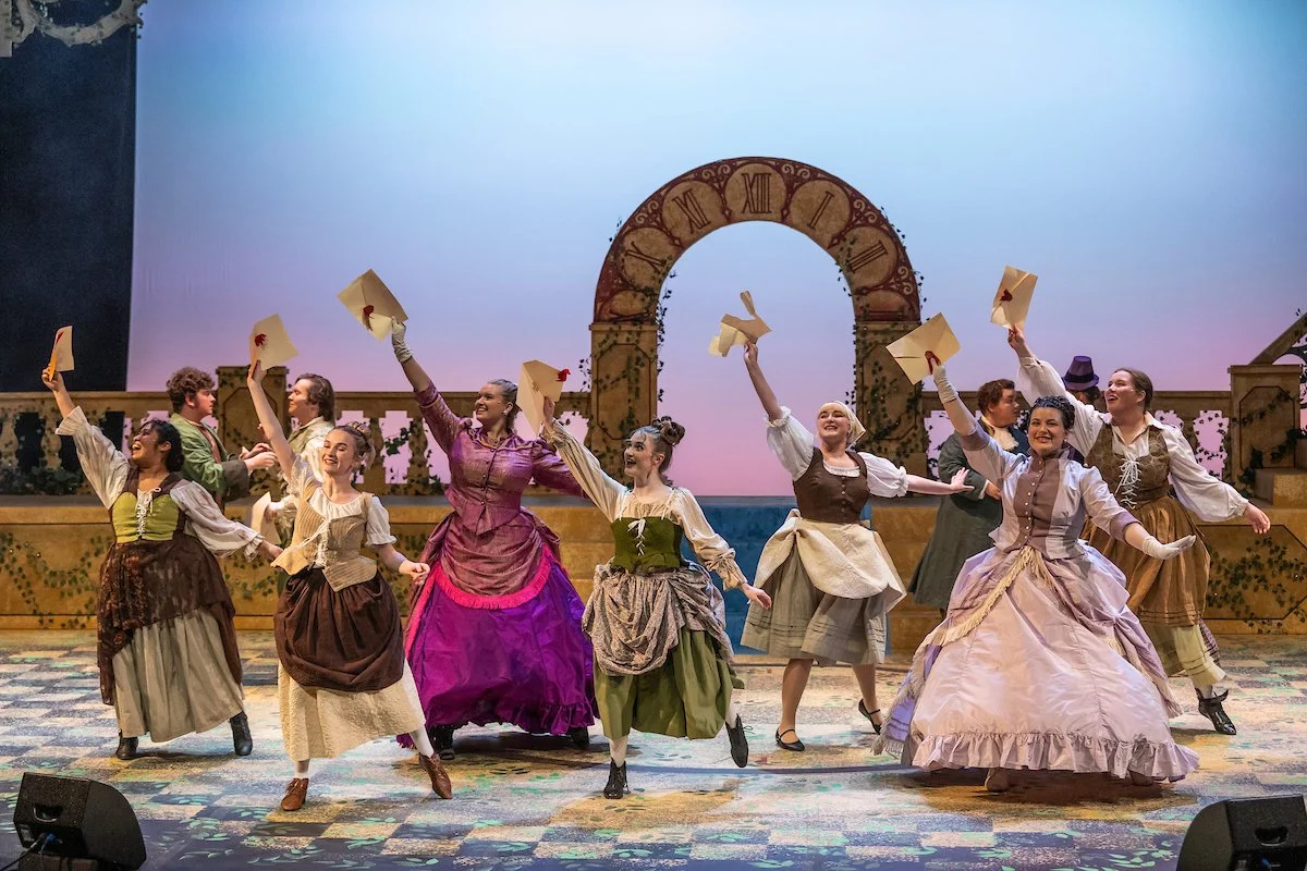 Theater performance with actors dressed in historical costumes, dancing on stage, with a backdrop featuring a clock arch and a painted sky.