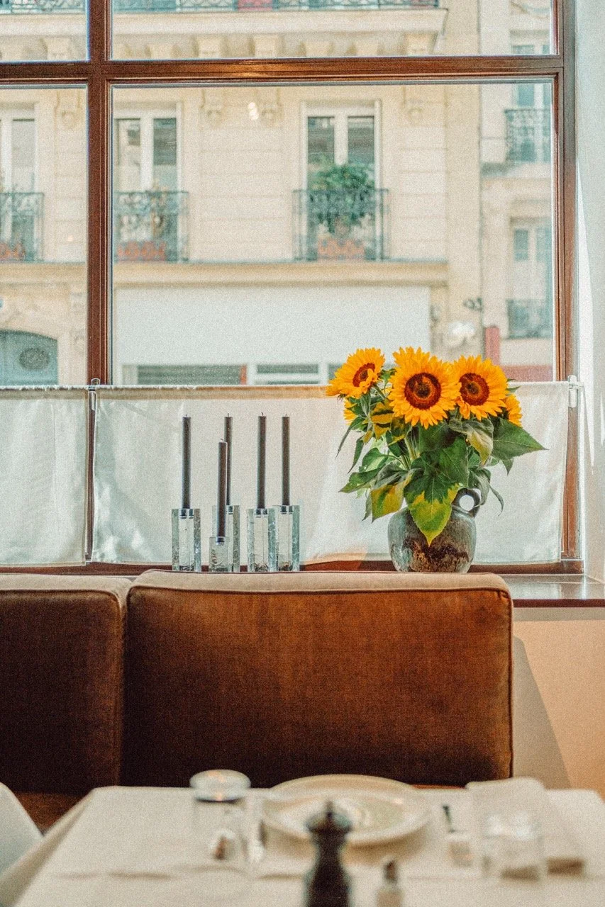 Sunflowers in a rustic vase on a windowsill with city buildings in the background, and some glasses on the windowsill, in a cozy restaurant or cafe setting.