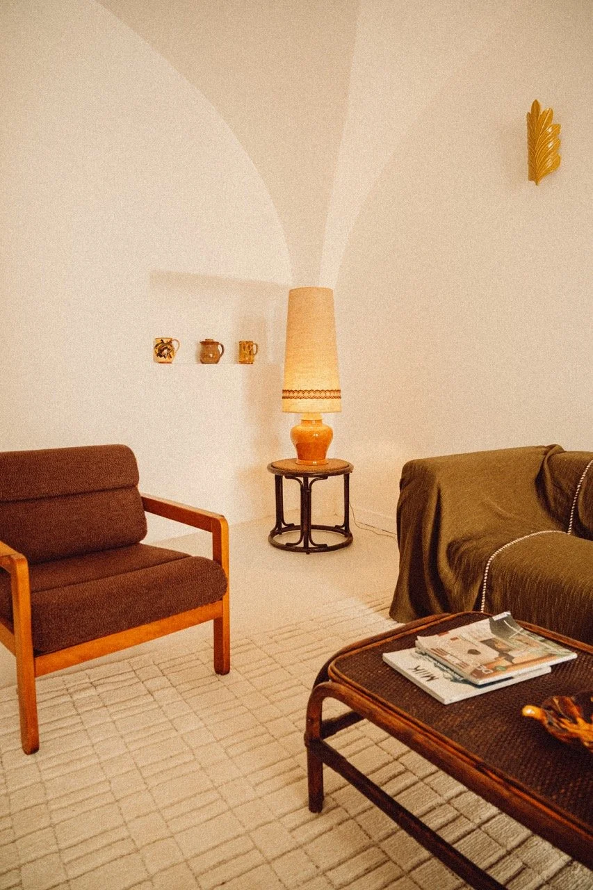 Living room with a brown armchair, a green sofa with a brown cover, a wooden coffee table with magazines, a side table with a large beige lamp, and three small decorative pots on a shelf built into the wall. There's a gold leaf wall decoration on the