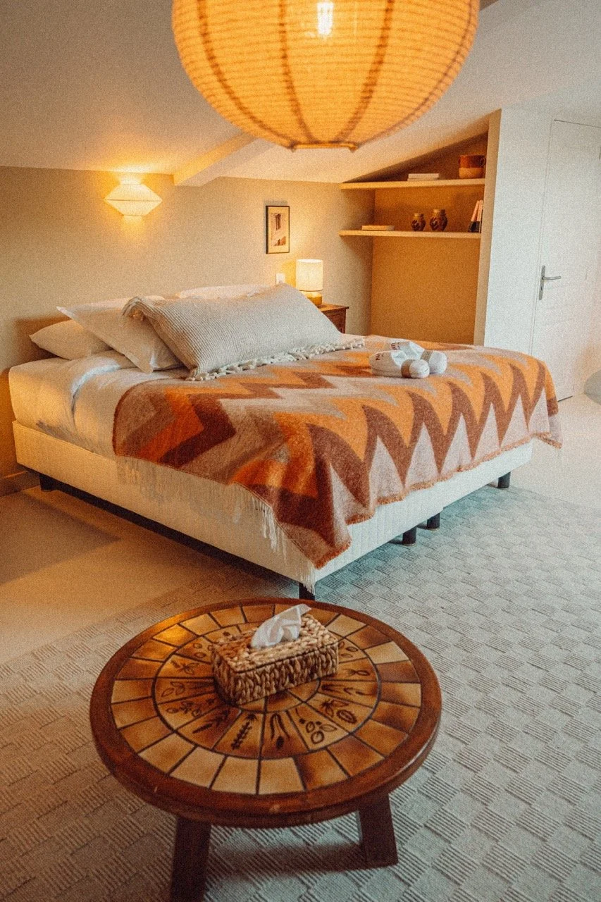 A cozy bedroom with a bed covered in an orange and beige geometric patterned blanket, decorative pillows, and a rolled towel; a small round wooden table with a tissue box and a clock design, and warm lighting from wall sconces and a table lamp.