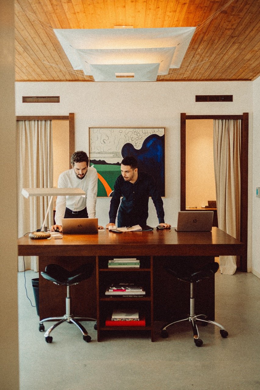 Two persons working at a wooden desk in a standing position with artwork hanging on the wall n the background