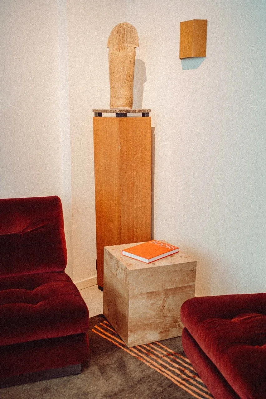 Living room corner with two red velvet chairs, a wooden pedestal with a stone sculpture on top, a small wooden cube table with an orange book, and a white wall with a small wooden wall hook.